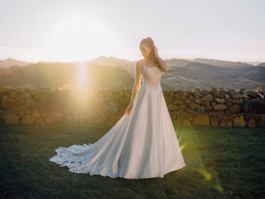 Bride in a flowing white wedding gown with a lace bodice and long train stands on grass at sunset beside a stone wall.