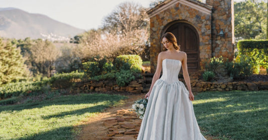 Bride wearing a full-length wedding dress stands on a stone path with a bouquet outside a rustic stone chapel.