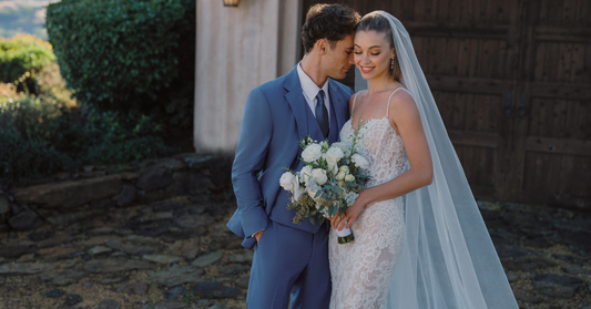 Bride in a lace white gown links arms with a groom in a blue suit as they lean their heads together outdoors.
